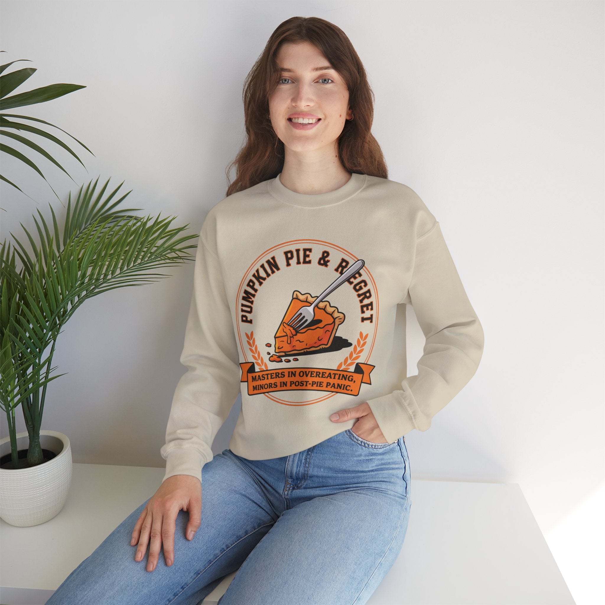 Woman wearing a Beige sweatshirt with a pumpkin pie graphic and text, sitting on a white surface with a plant in the background.
