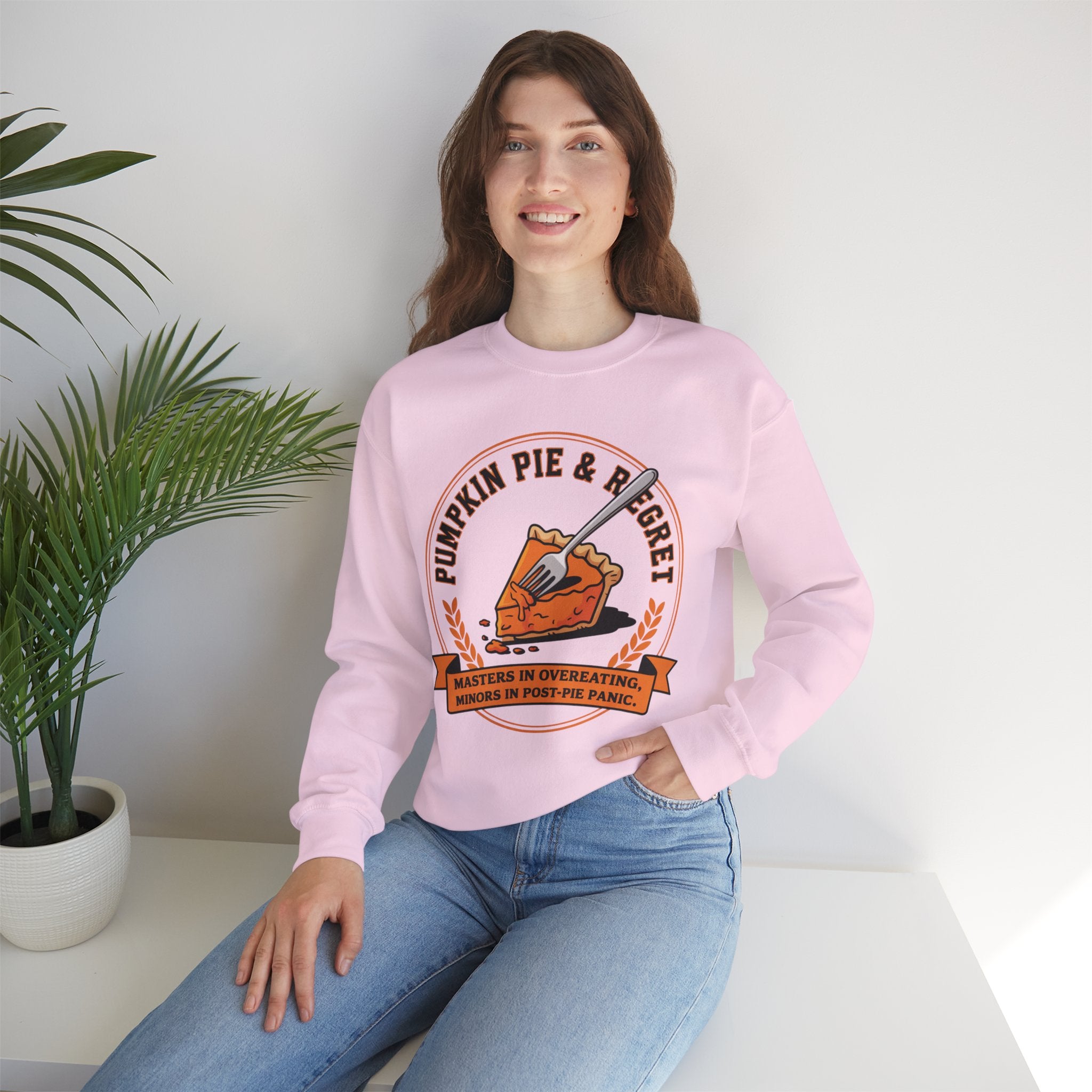 Person wearing a Light Pink sweatshirt with a pumpkin pie design and text, sitting on a white surface with a plant in the background.