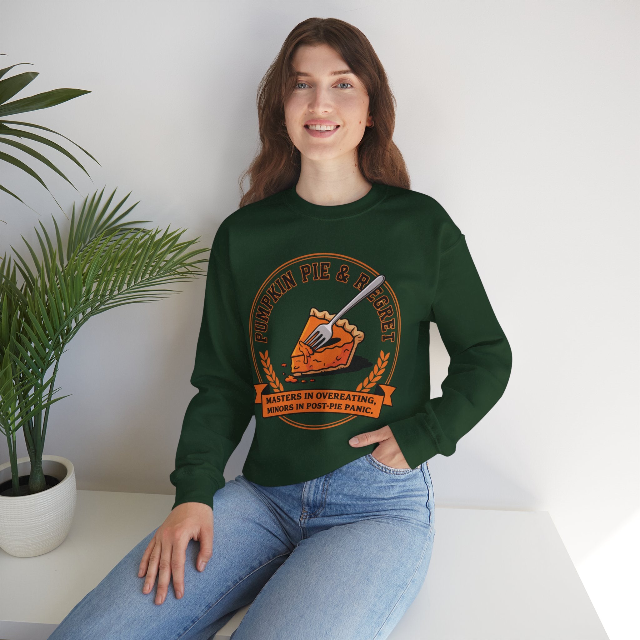 Woman wearing a Forest Green sweatshirt with a pumpkin pie graphic design, sitting on a white surface with a plant in the background.